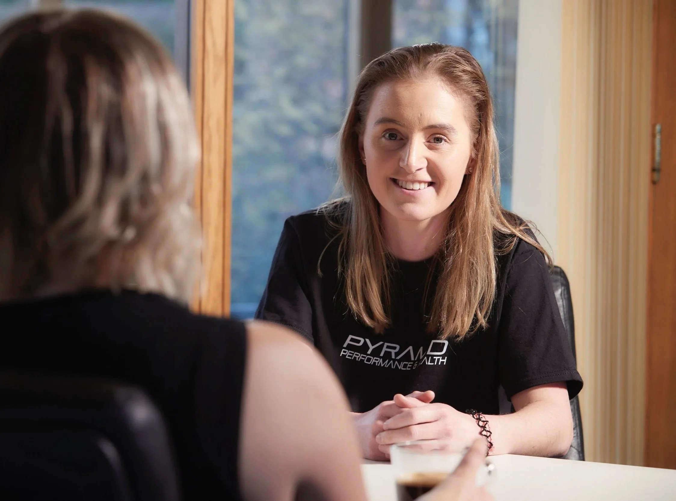 A woman with long brown hair smiling and wearing a black T-shirt with white text, sitting at a table and engaging in conversation with another person. The background includes a window with a view of trees.