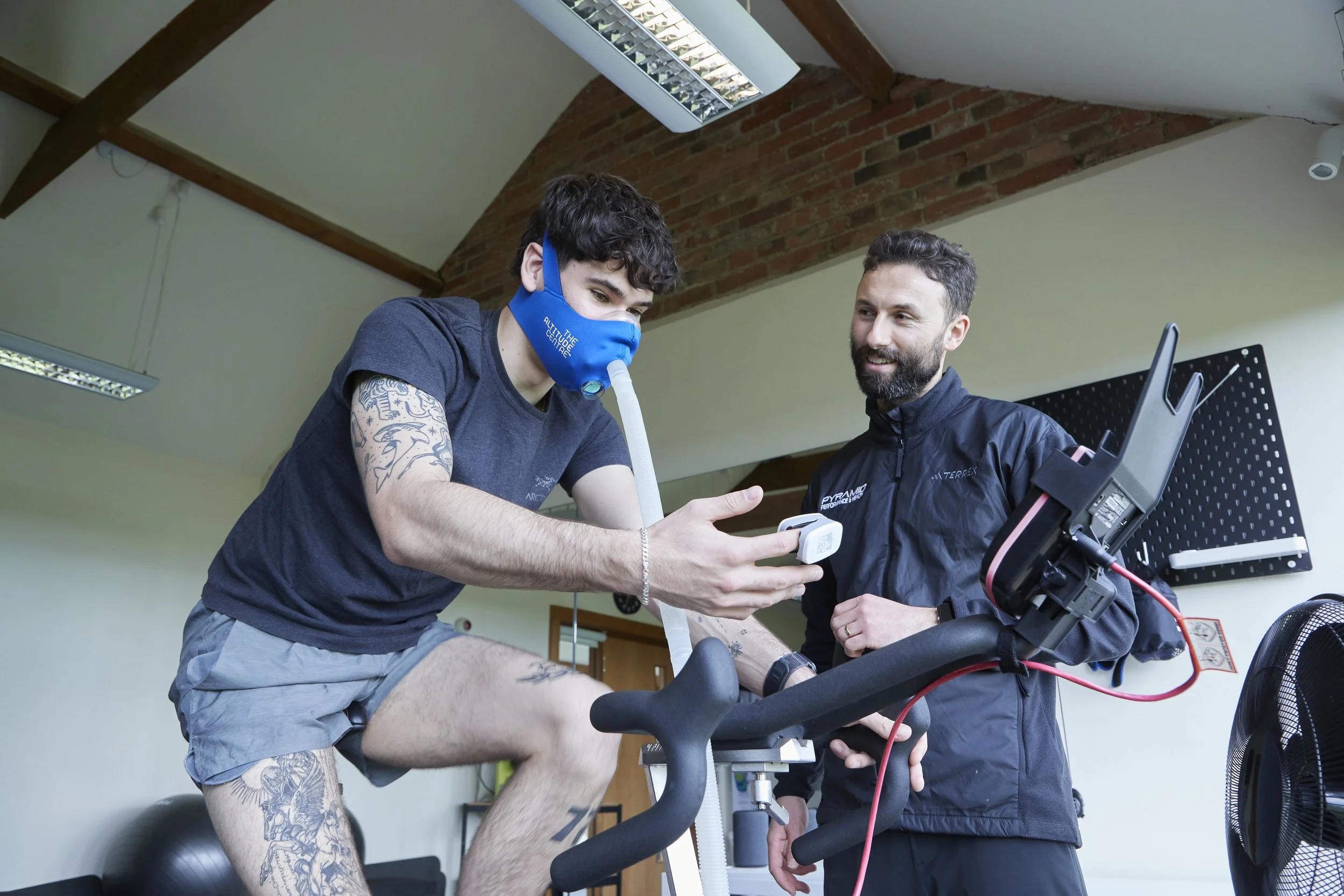 A man in workout shorts and tattoos on his arm undergoes a fitness test on a stationary bike while a trainer monitors him with a computer and equipment in a gym or medical setting.