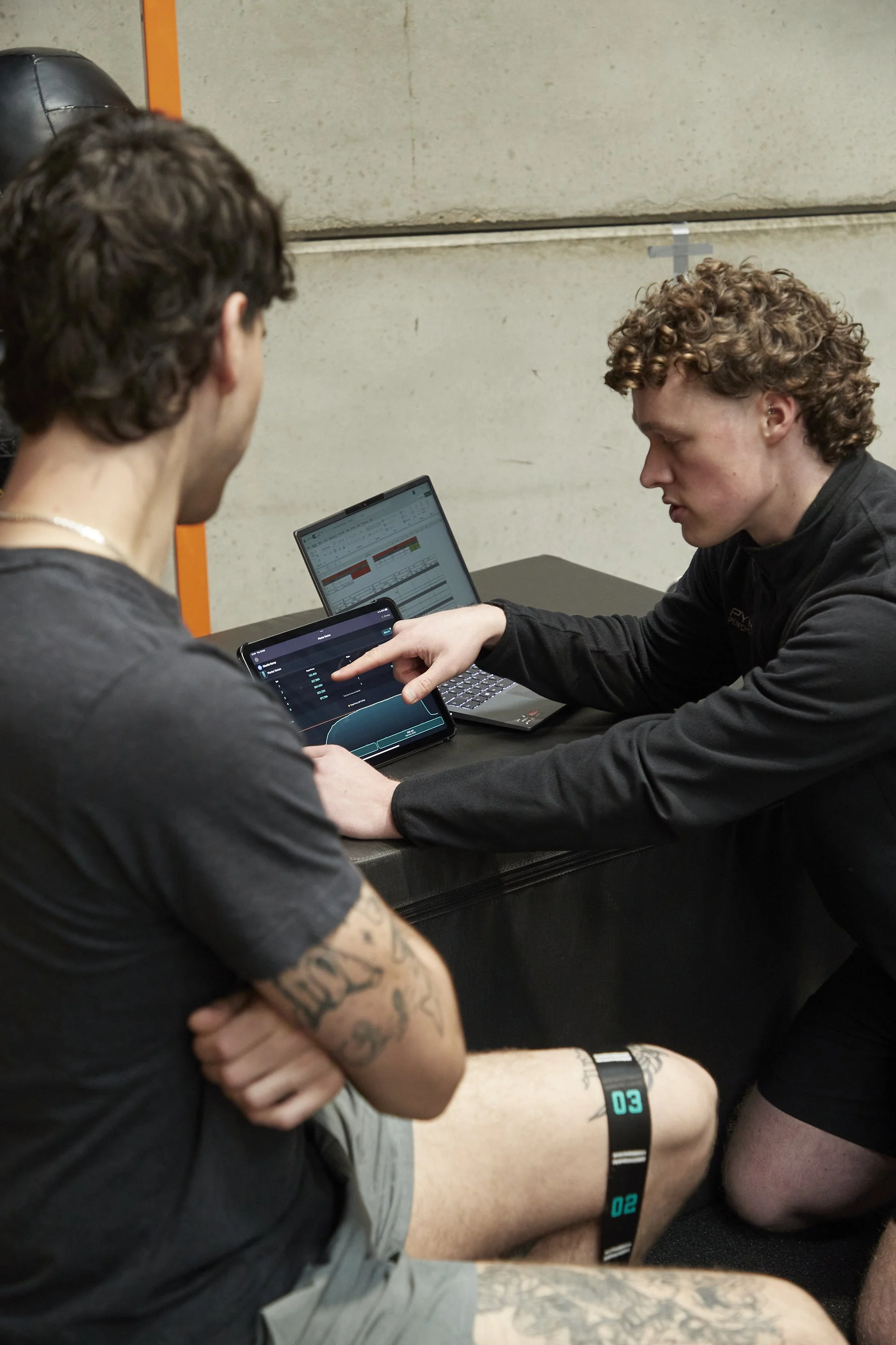 Two young men sitting at a table with laptops and tablets, engaged in a discussion. One man has tattoos and is wearing a black shirt, the other has curly hair and is wearing a black jacket. The setting appears to be an indoor workspace or meeting are