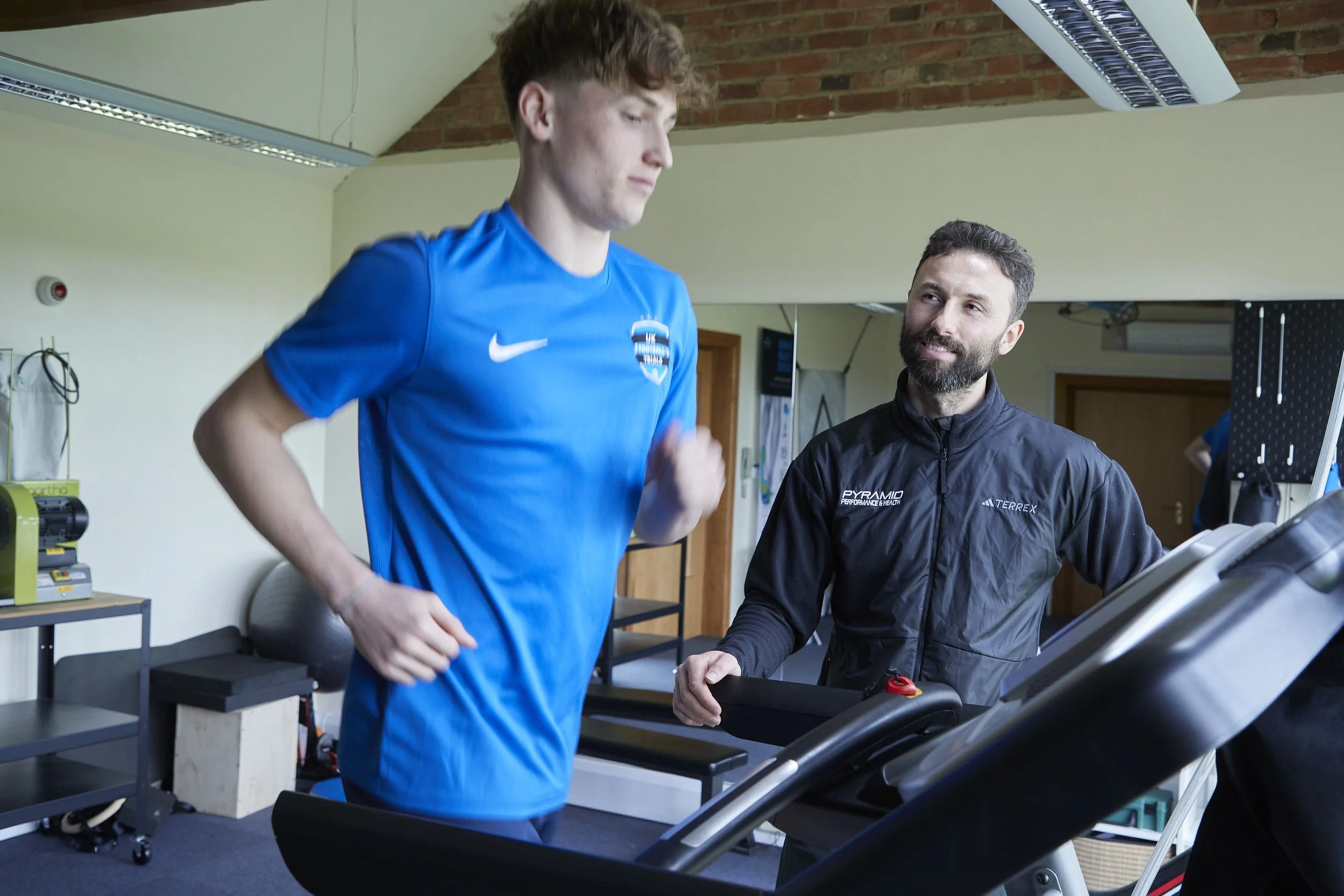 A young man in a blue sports shirt jogging on a treadmill while talking to a trainer in a black jacket with white text, inside a fitness center with exposed brick walls and gym equipment.