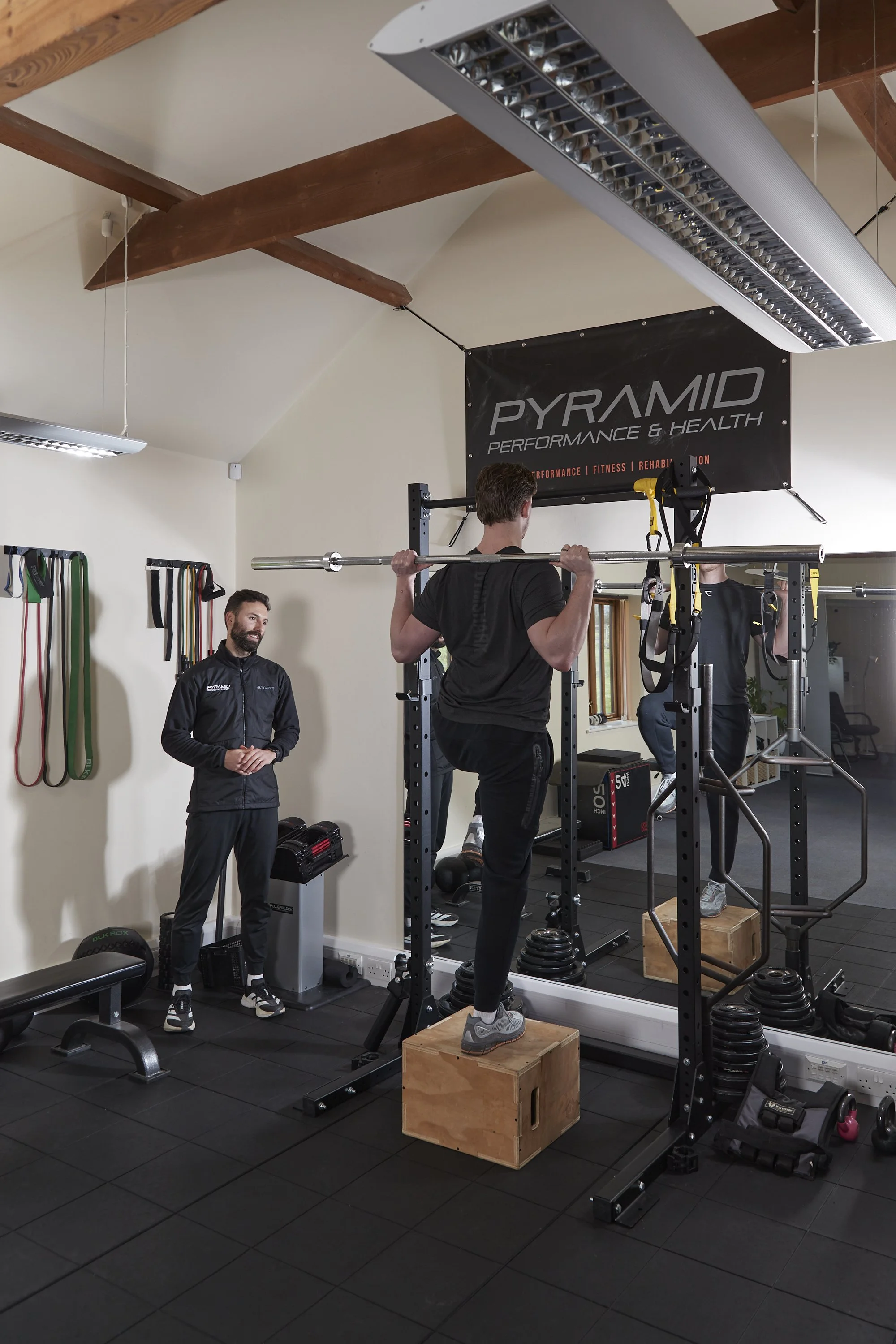 People in a gym performing weightlifting exercises with a weight rack, weights, and fitness equipment visible, with a trainer overseeing.