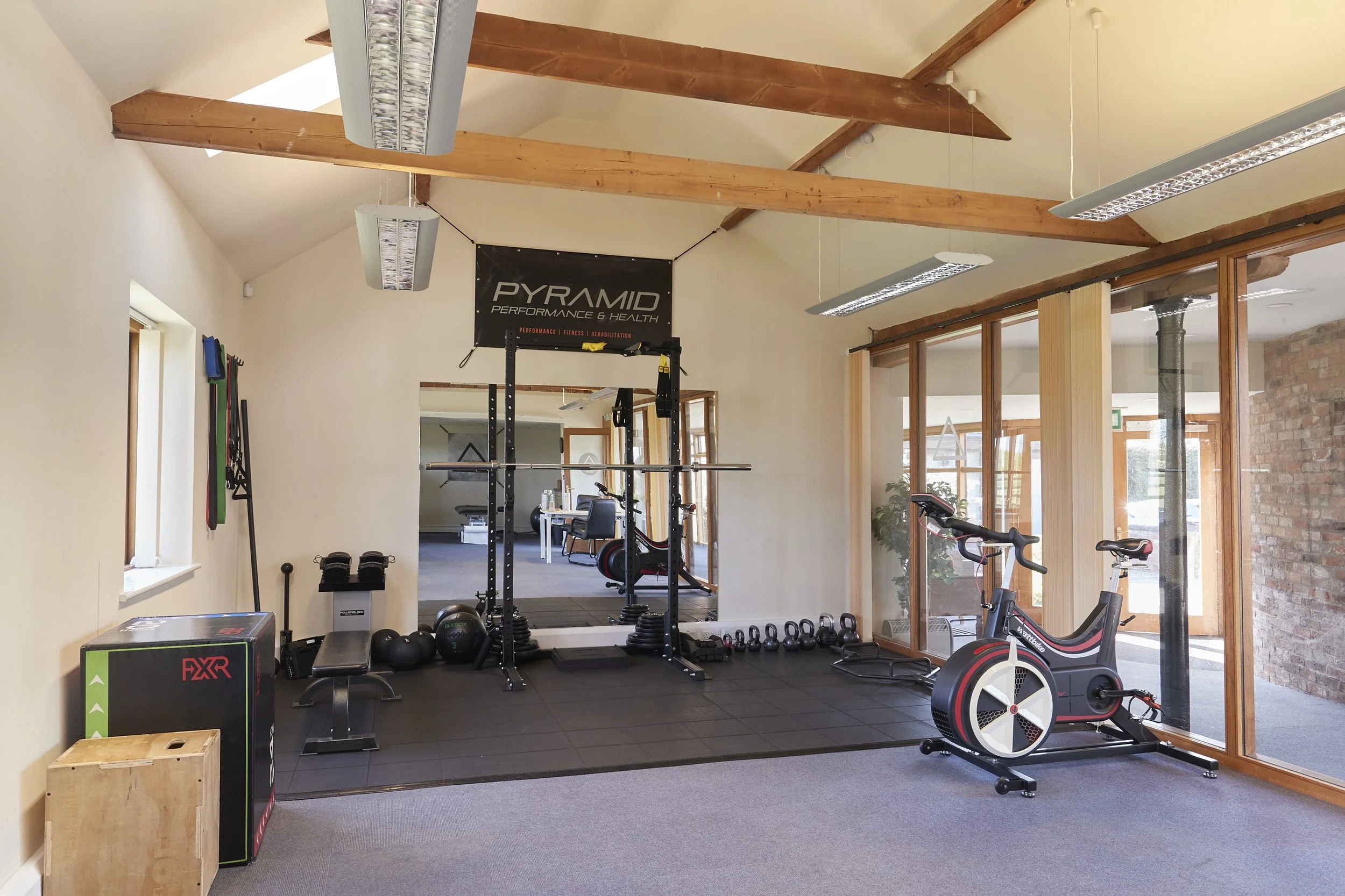 Indoor gym with wooden beams on the ceiling, various exercise equipment including squat rack, exercise bike, weights, and medicine balls, and large windows letting in natural light.