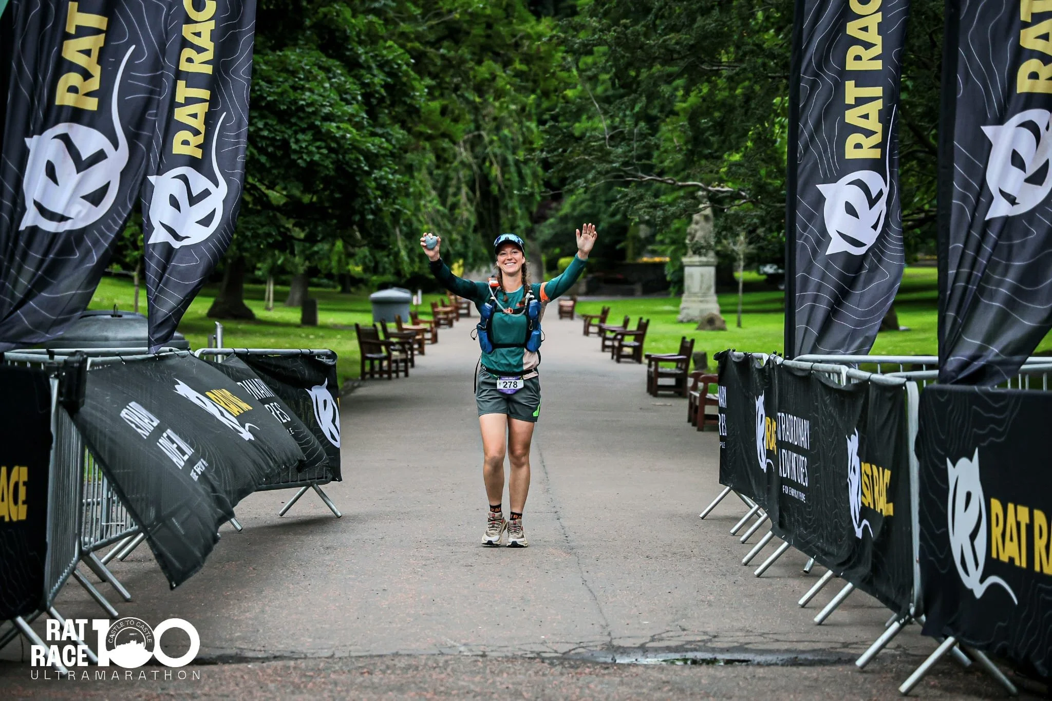 A female runner smiling as she reaches the finish line of a race in a park. She is wearing a teal jacket, shorts, and a cap, with her arms raised in celebration. The finish line is marked with black banners displaying "RATRACE" and other race info, and there are green trees and benches in the background.