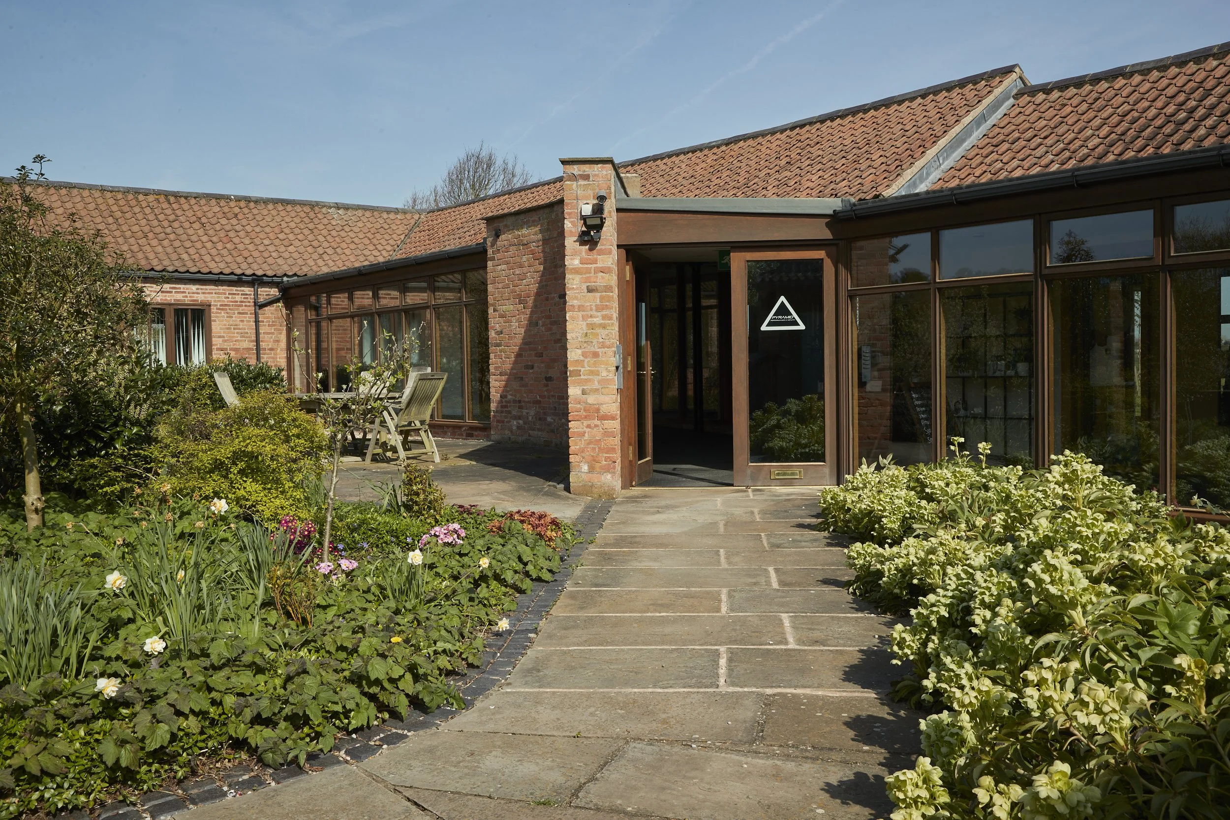 Sunlit garden with stone pathway leading to a brick and glass building with a tiled roof and outdoor seating.