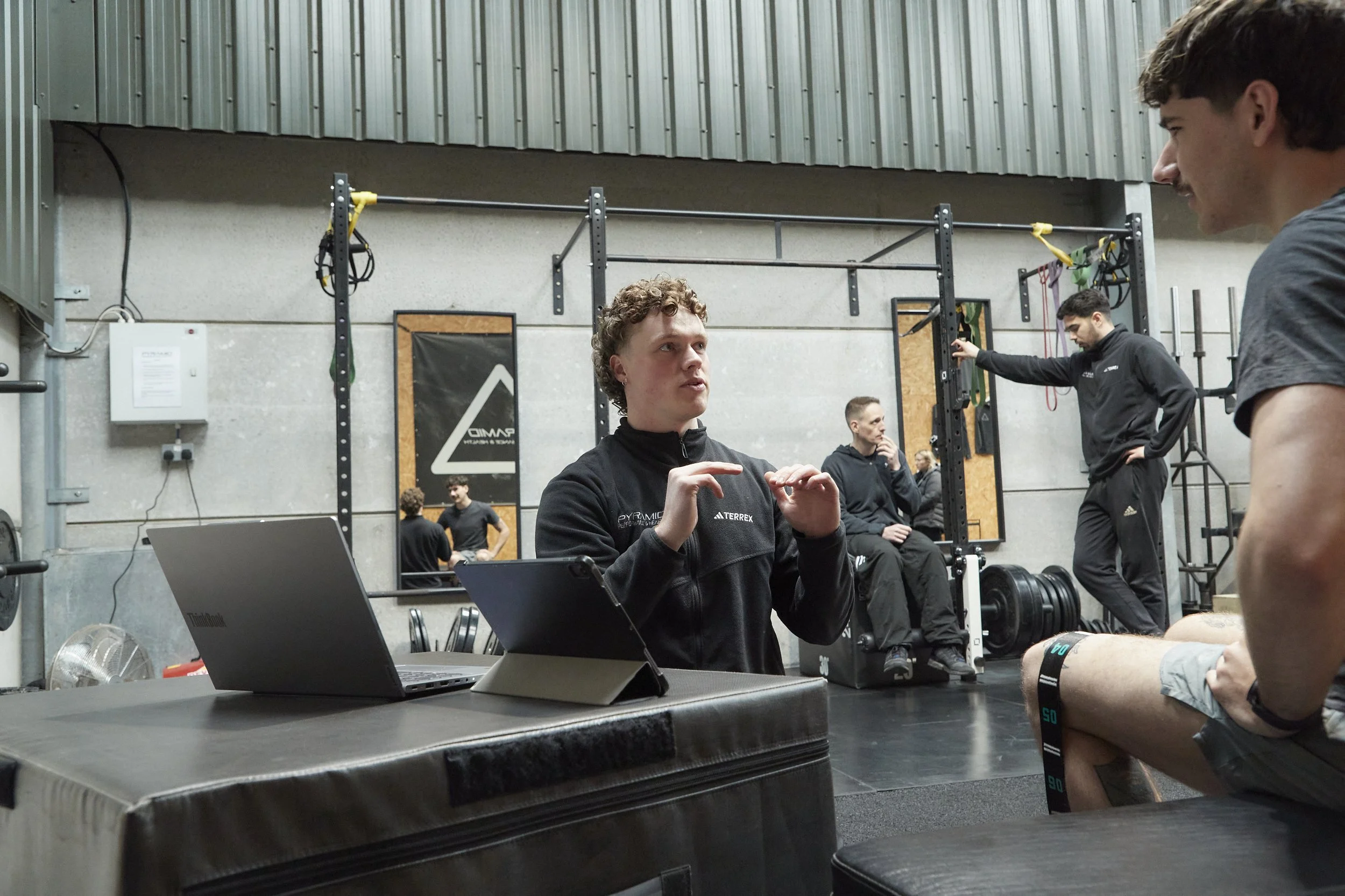 Three men in athletic clothing inside a gym, with one sitting at a table with a laptop, talking to another who is standing. In the background, two more men are near workout equipment, one adjusting a machine and another sitting on a bench. The gym ha