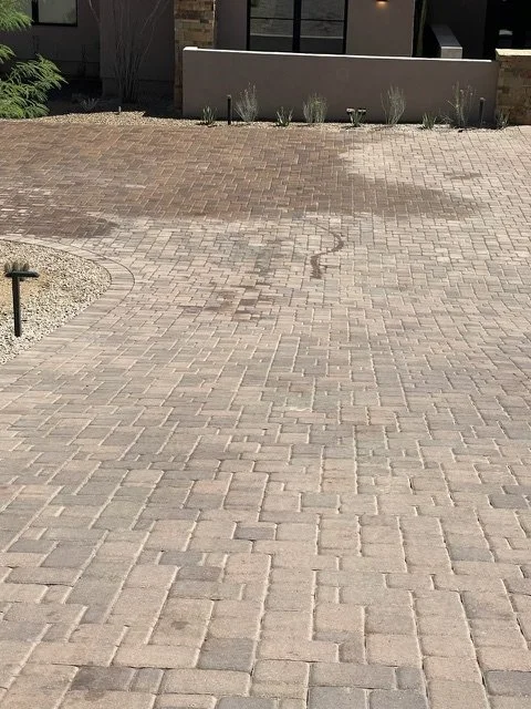 A paved outdoor area with light brown bricks, some arranged in a herringbone pattern, and a small landscaped section with plants and gravel, with a modern building in the background.