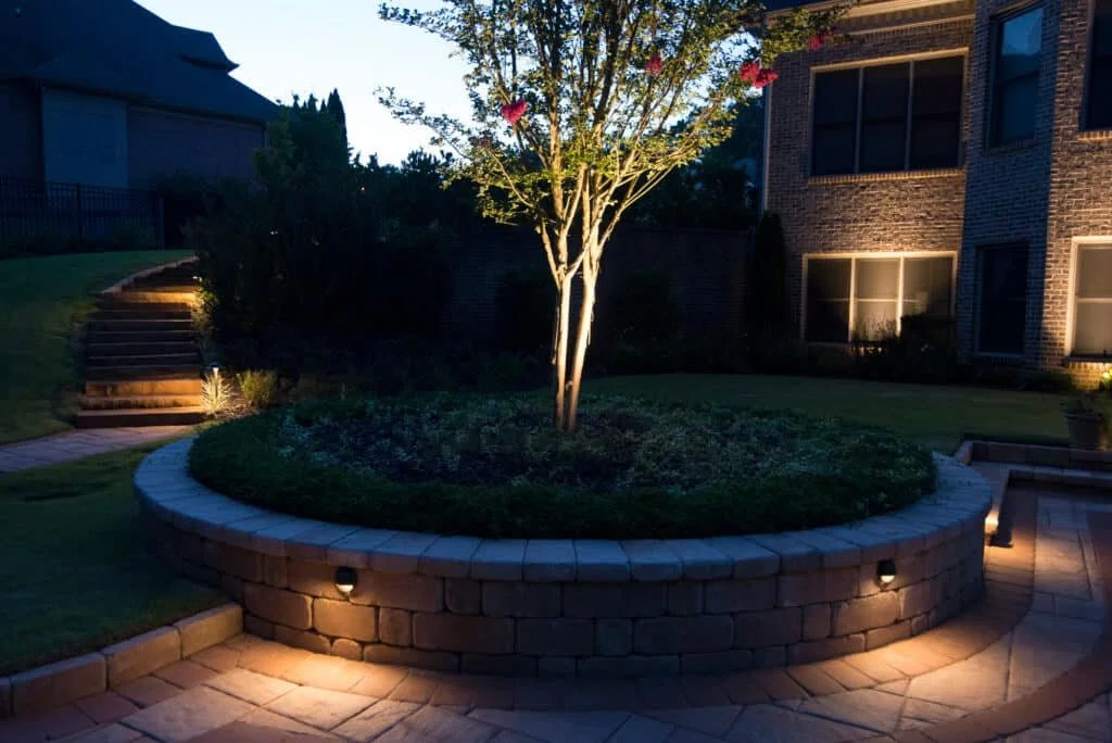 Nighttime view of a backyard with a brick planter containing a tree, surrounded by a brick patio illuminated by ground lights, with steps on the left leading to a grassy area and neighboring houses.