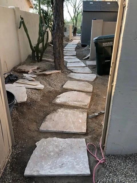 Outdoor backyard with a stone pathway under construction, with large flat stone slabs laid on dirt, leading towards a tree and a storage shed in the background.