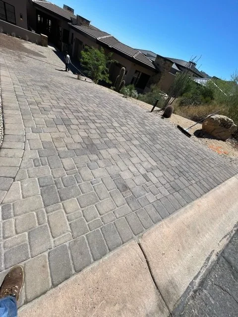 A paved sidewalk next to a curb with desert vegetation and rocks in the background. Residential buildings are visible in the distance under a clear blue sky.