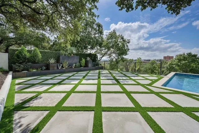 Modern backyard with a geometric concrete and grass patio, a swimming pool on the right, and lush greenery and trees in the background under a partly cloudy sky.