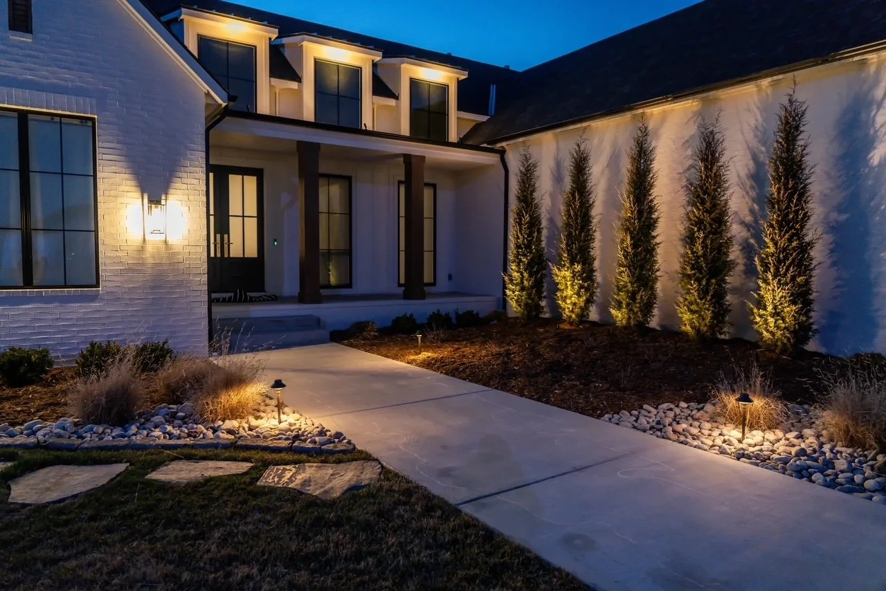 Nighttime view of a modern house with exterior lighting, a concrete walkway, tall narrow trees along a white fence, and landscaped yard with rocks and small illuminated plants.