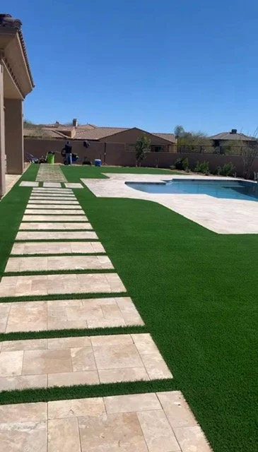 A backyard with a rectangular swimming pool, beige stone tiles surrounding the pool, a green artificial grass lawn, and a stone pathway leading to a sitting area with chairs under a clear blue sky.