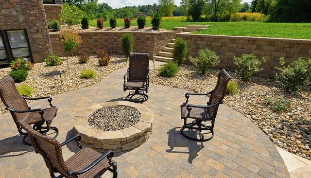 Outdoor patio with four rocking chairs arranged around a stone fire pit, surrounded by landscaping and stairs leading up to a grassy area.