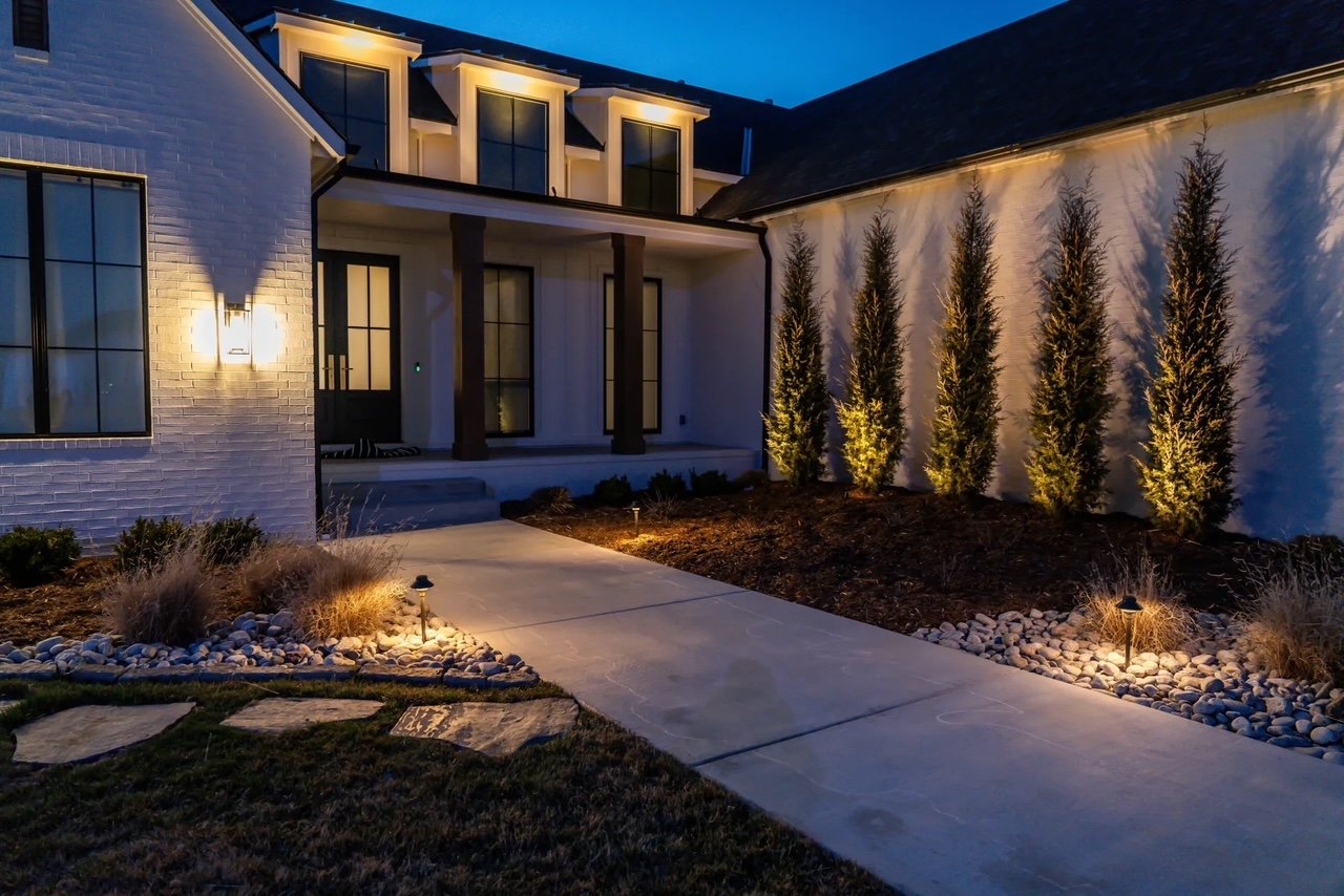 Night view of a modern house with front porch, illuminated outdoor wall lights, concrete walkway, tall evergreen trees along side yard, decorative rocks, and landscape lighting.