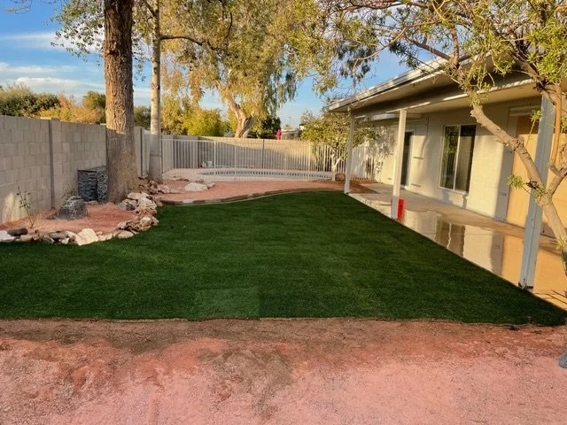 Backyard with a lawn, trees, and a house with a patio. The yard is partially landscaped with rocks and a concrete path.