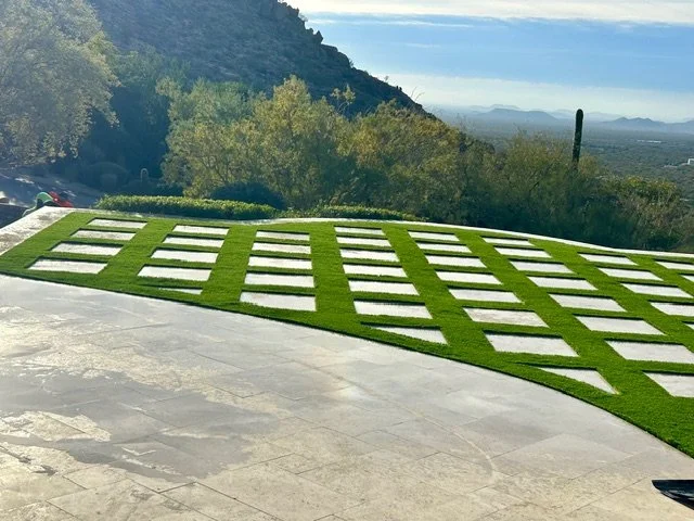A modern outdoor area with a concrete patio and a decorative grass-covered section arranged in a geometric pattern, overlooking a landscape with trees and mountains in the distance.
