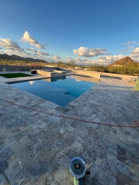 In-ground swimming pool with gray stone patio, overlooking mountains and blue sky with clouds, on a sunny day, with pool equipment nearby.