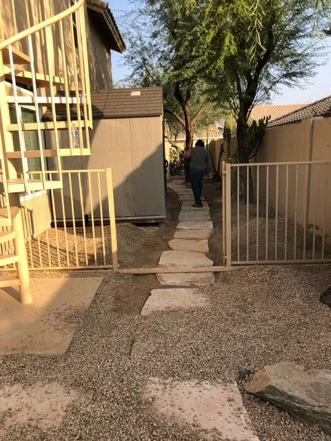 Person walking down a small stone pathway between beige fences with trees and houses in the background.