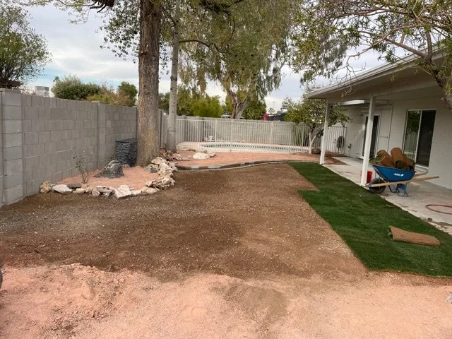 Backyard with partially laid sod grass, construction materials, and tools. Backyard is enclosed with a brick wall and a white wooden fence, with a house on the right side.