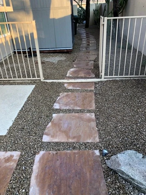 Stone pathway leading through a white metal gate, with gravel on either side and a small garden area to the right.