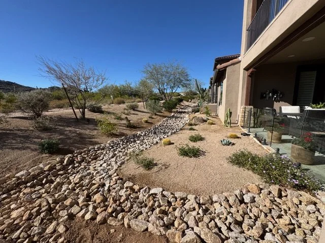 Desert landscape with rocks, small bushes, and a few dry trees, near a residential house with a balcony and outdoor patio on a sunny day.
