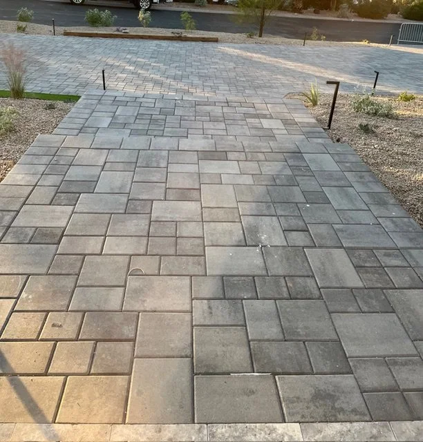 Concrete paver walkway leading to a parking lot with a few small plants and low landscaping borders.