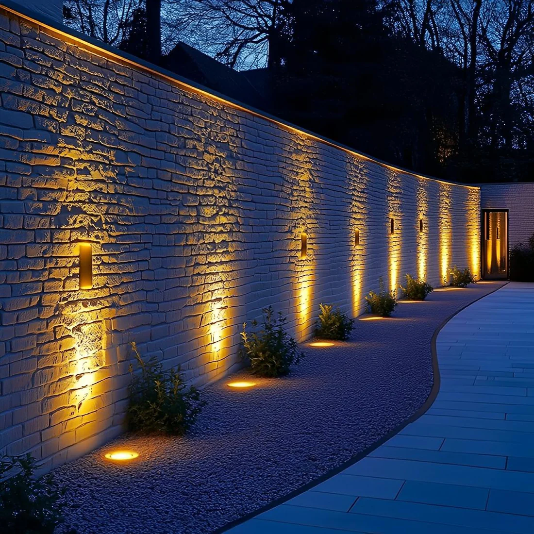 A modern exterior wall illuminated by warm yellow lights at night. The wall is made of white bricks with garden plants at the base and a paved pathway in front.