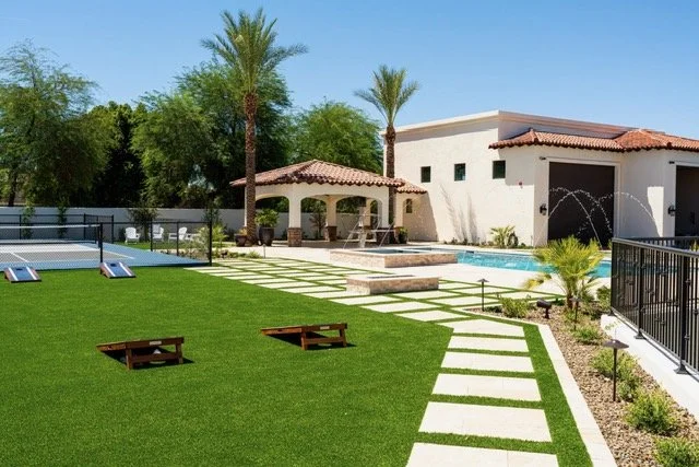 Luxury backyard with a well-manicured lawn, palm trees, a swimming pool, and a modern white house with a red-tile roof.
