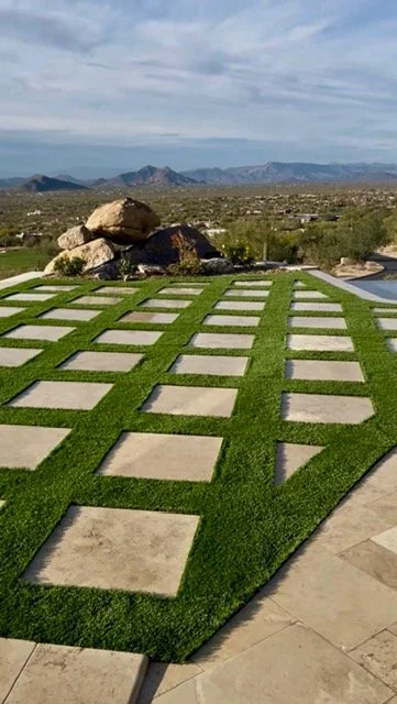 Modern outdoor space with a patterned lawn featuring square concrete pavers surrounded by green grass. In the background, there are large rocks and a view of distant mountains under a cloudy sky.
