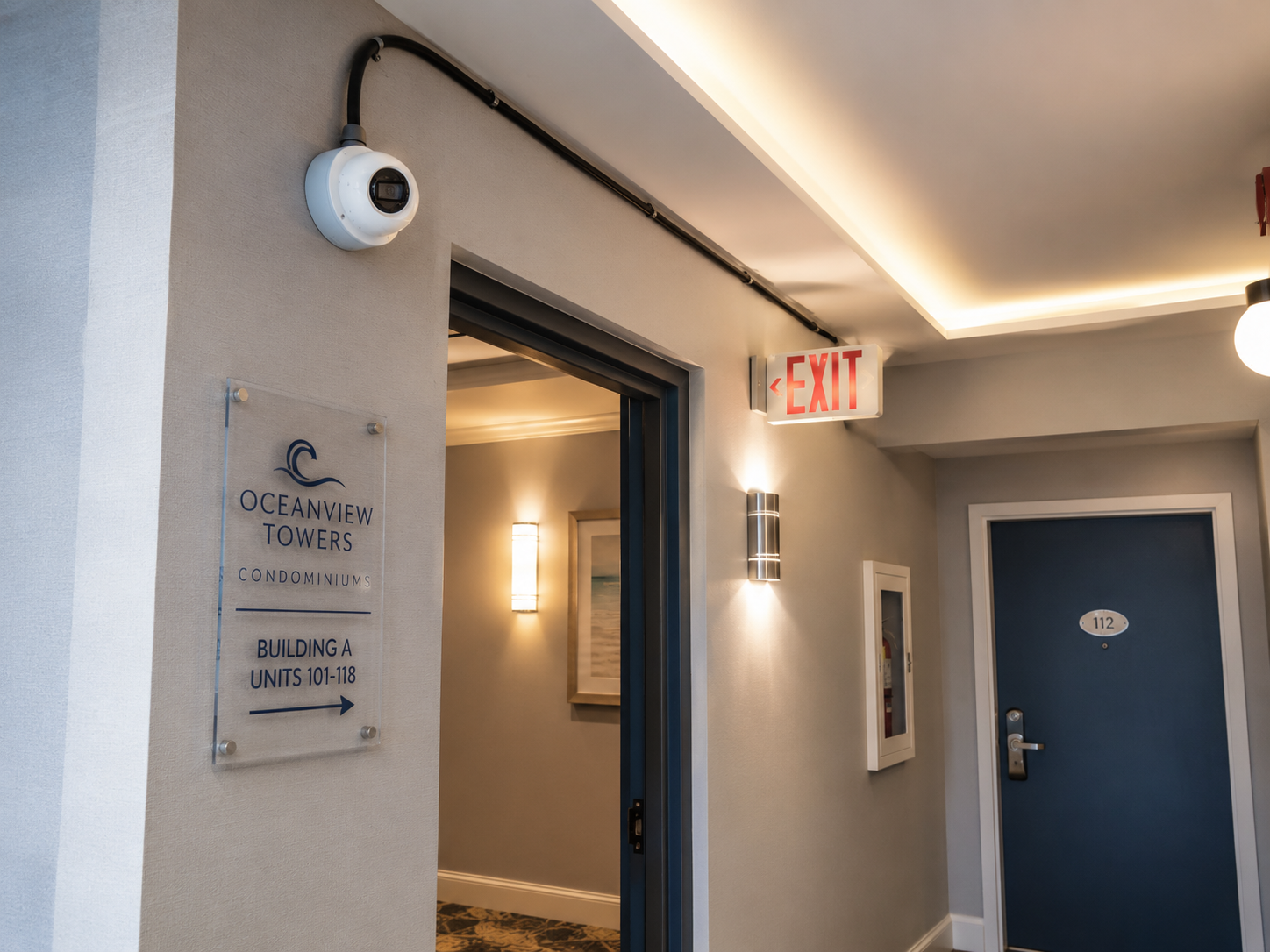 Hallway with exit sign, security camera, Oceanview Towers condominium sign, door labeled 112, and interior lighting.