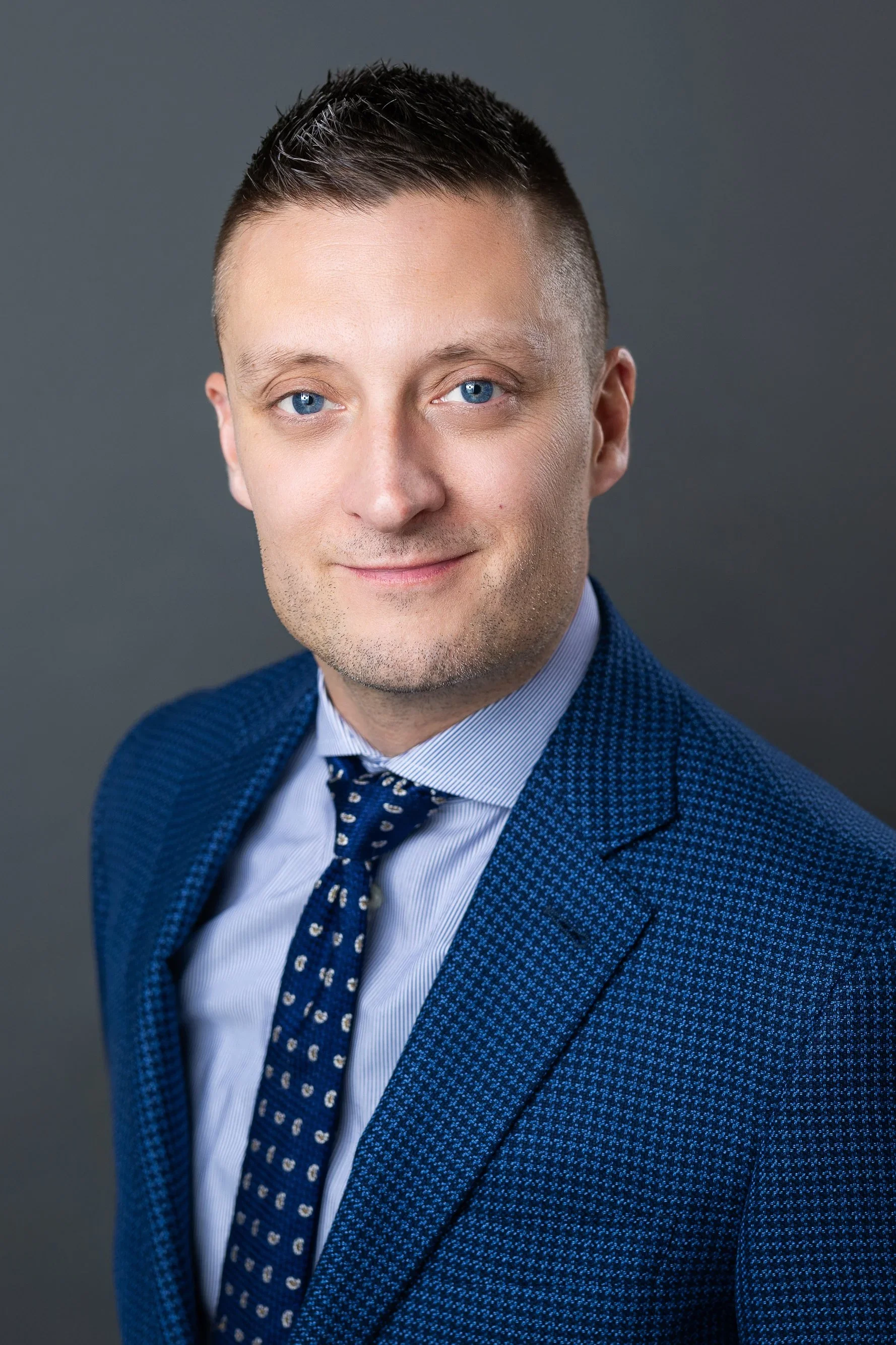 A man with short dark hair, blue eyes, light skin, wearing a blue textured blazer, a light blue shirt, and a navy blue tie with a pattern, against a gray background.