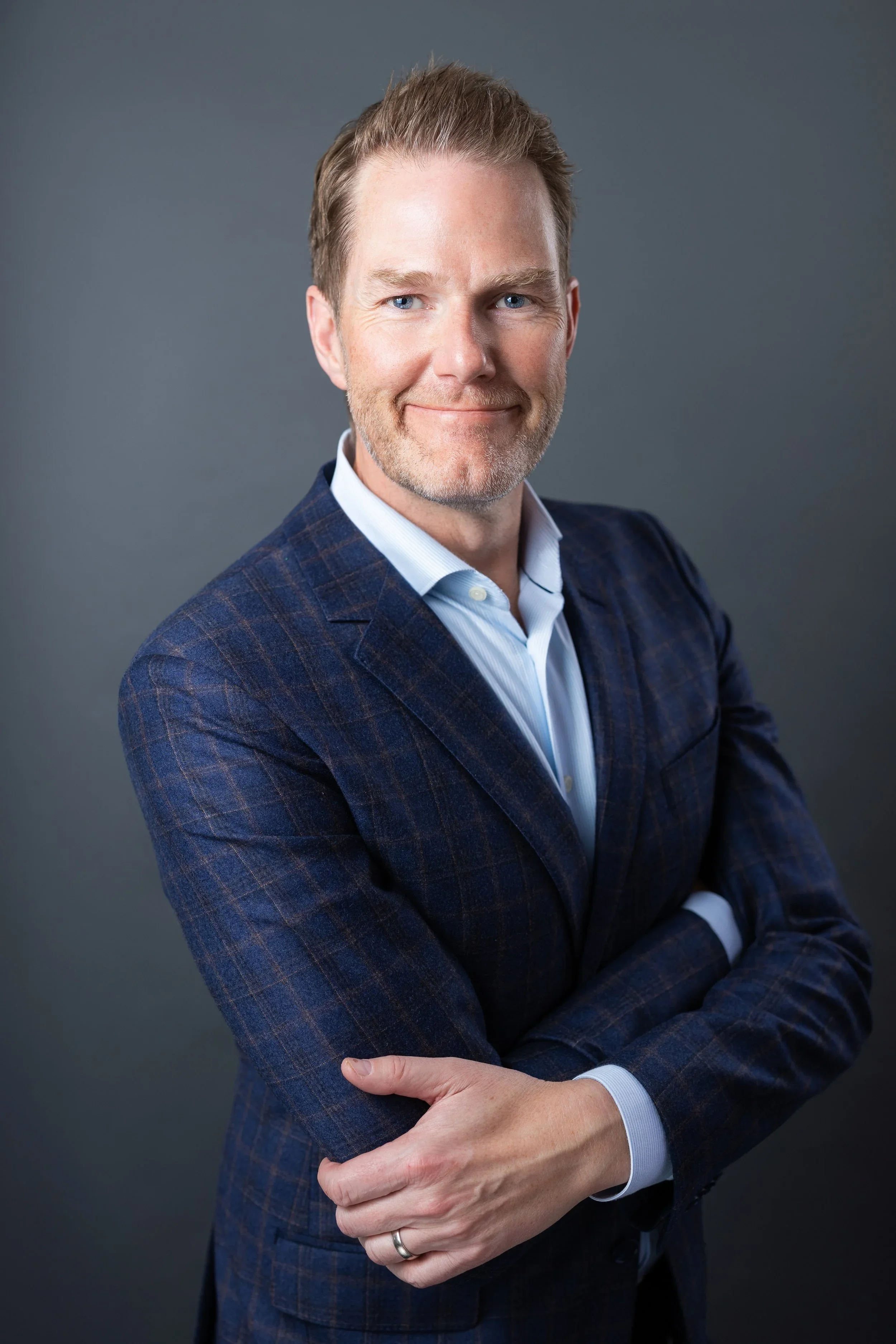 A man with light skin, light brown hair, and a beard, smiling and posing with arms crossed, wearing a dark blue plaid blazer and a light blue dress shirt, against a dark gray background.