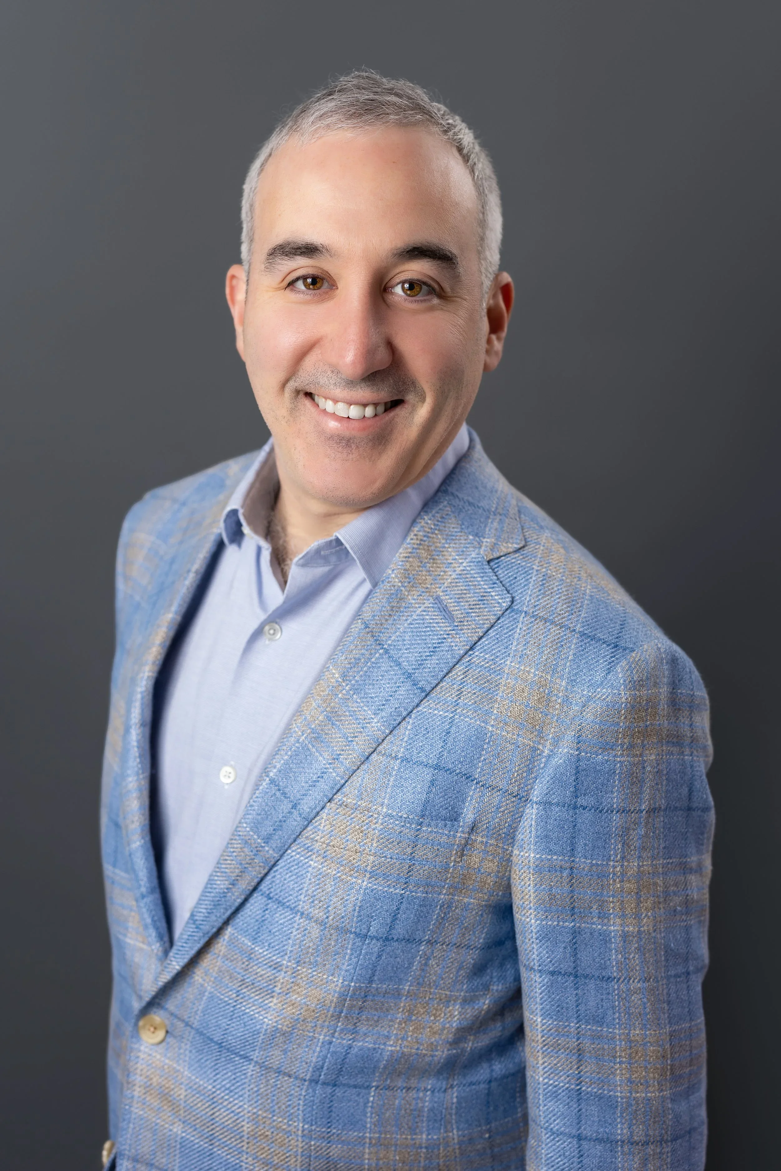 A man with short gray hair, smiling, wearing a light blue dress shirt and a checked blazer against a dark gray background.