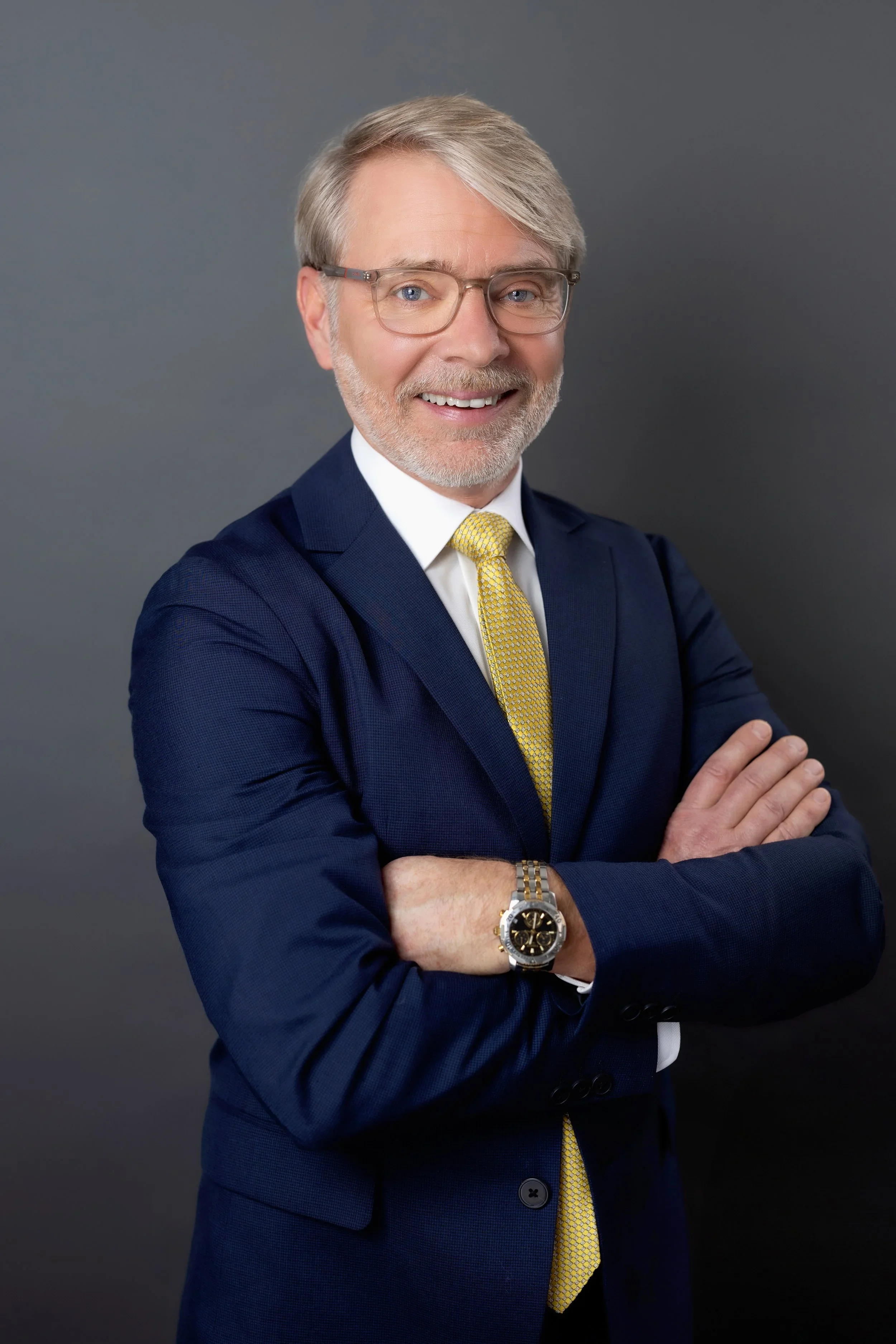 A man with gray hair and beard smiling, wearing glasses, a dark blue suit, a white shirt, a yellow tie, and a silver wristwatch, standing with crossed arms against a gray background.