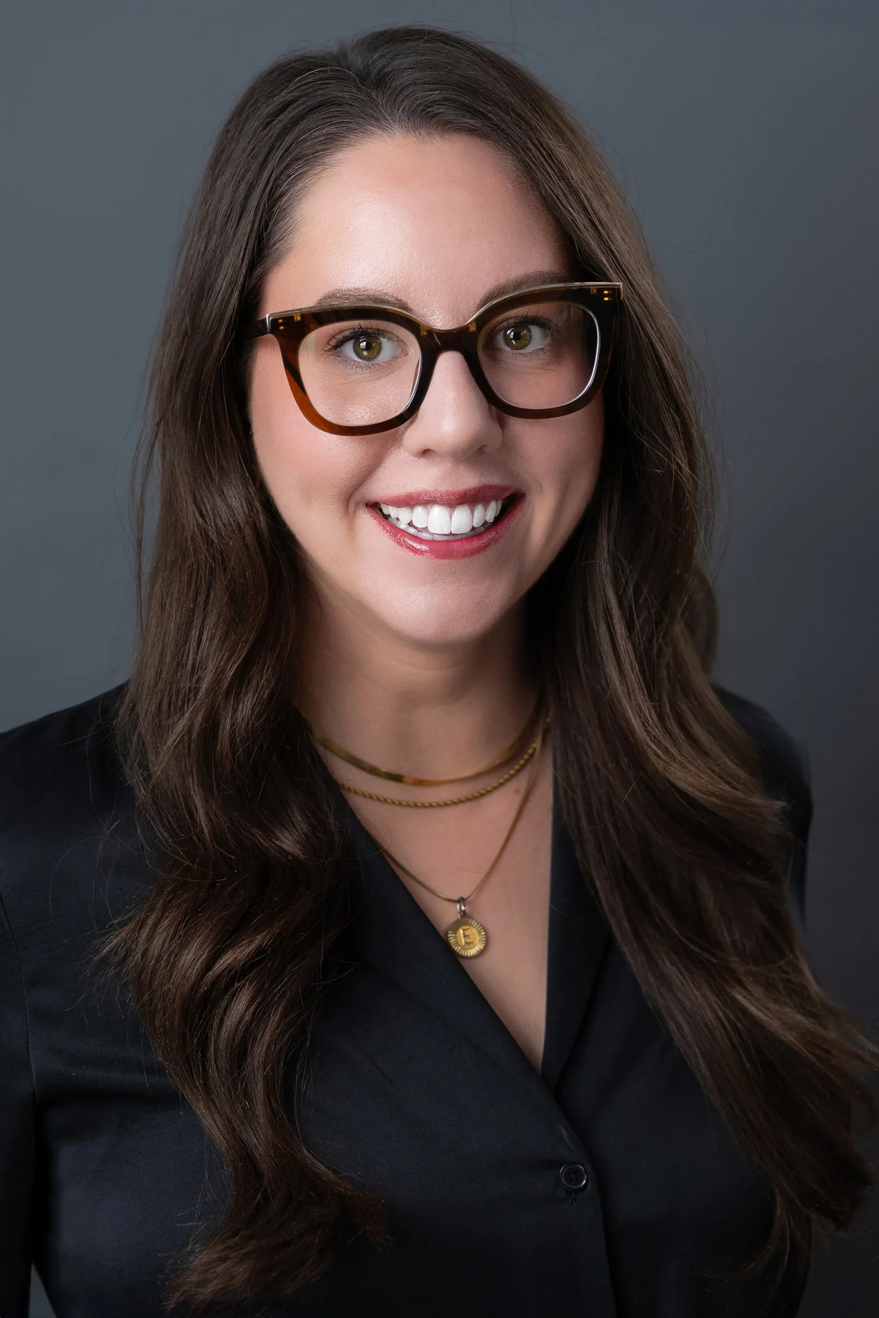 A woman with long brown hair wearing large, tortoise-shell glasses, a black blazer, and gold jewelry, smiling against a gray background.