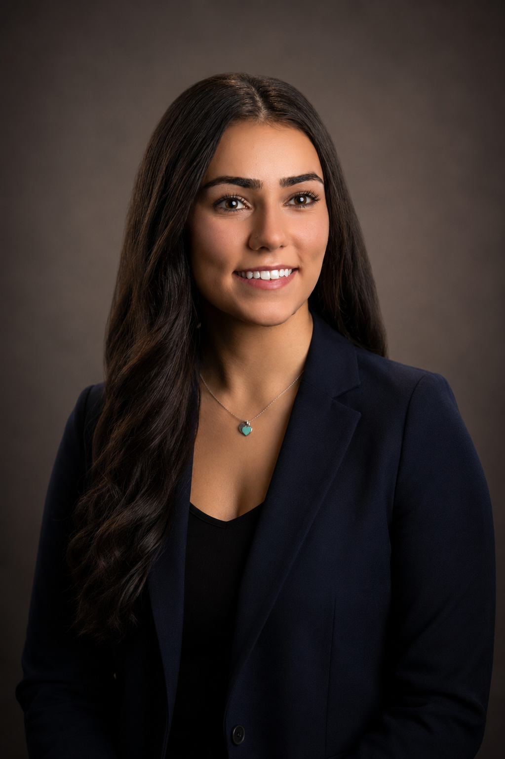 Professional portrait of a young woman with long dark hair, wearing a black top, navy blazer, against a dark grey background.