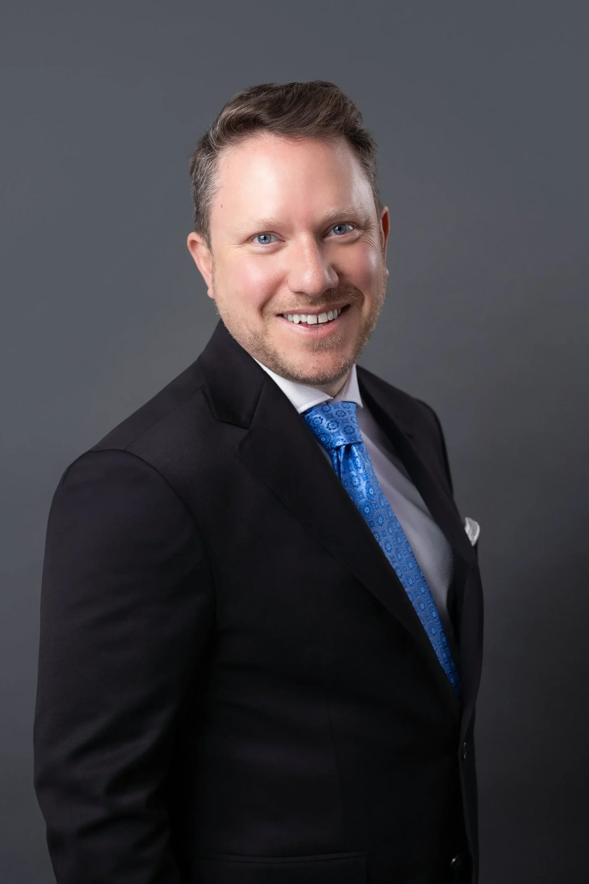 A smiling man in a black suit, white shirt, and a blue patterned tie against a solid gray background.
