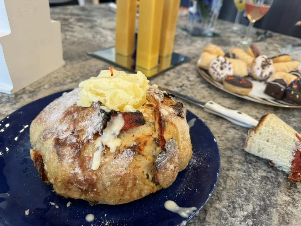 A plate with a large, round, breaded meatball topped with mashed potatoes and drizzled with white gravy, on a dark blue plate. In the background, there is a plate of assorted cookies and pastries, a knife, two glasses of rosé wine, and a brown paper bag on a speckled granite countertop.