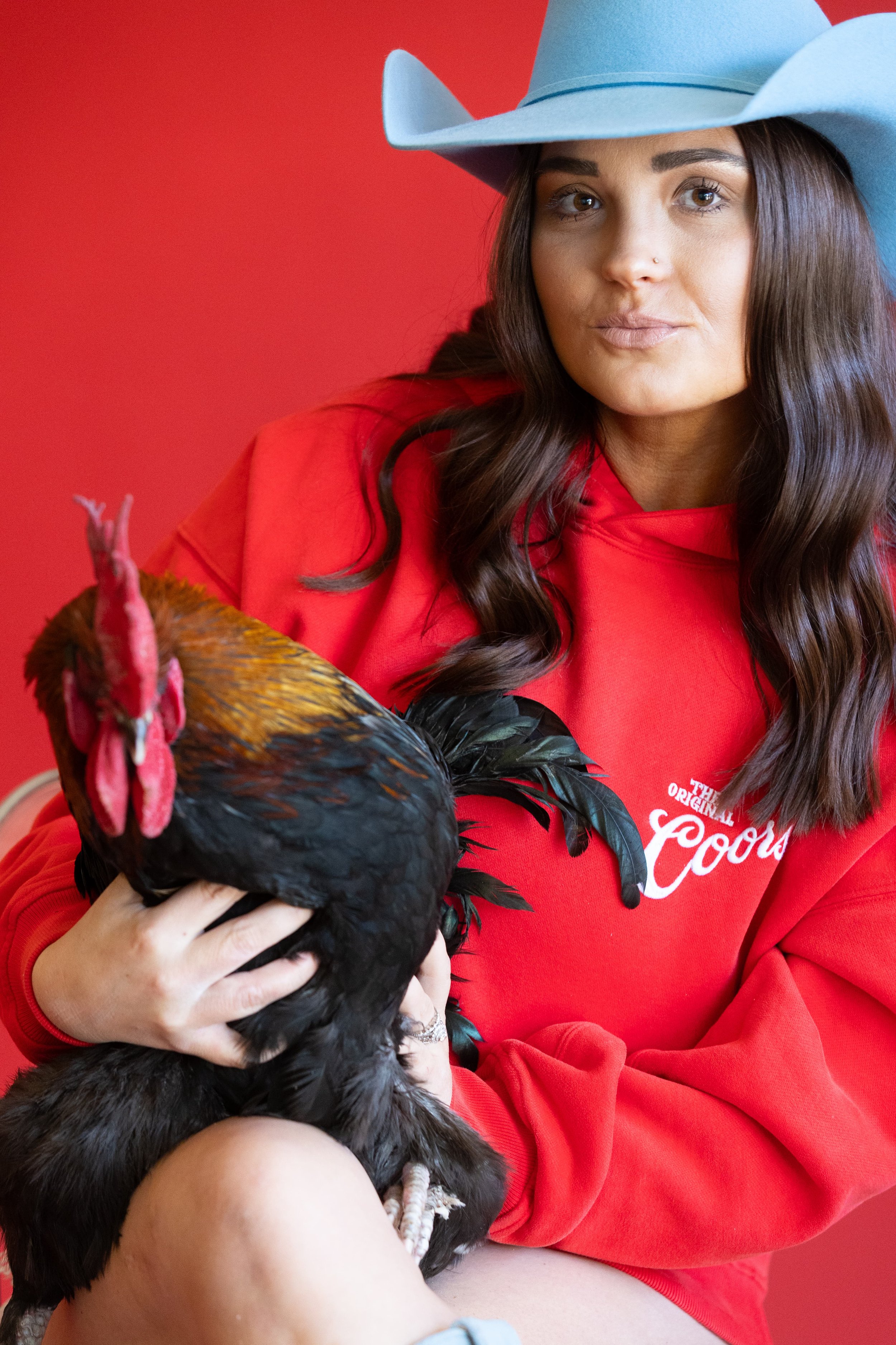 A woman wearing a blue cowboy hat and red sweatshirt holding a rooster against a red background.