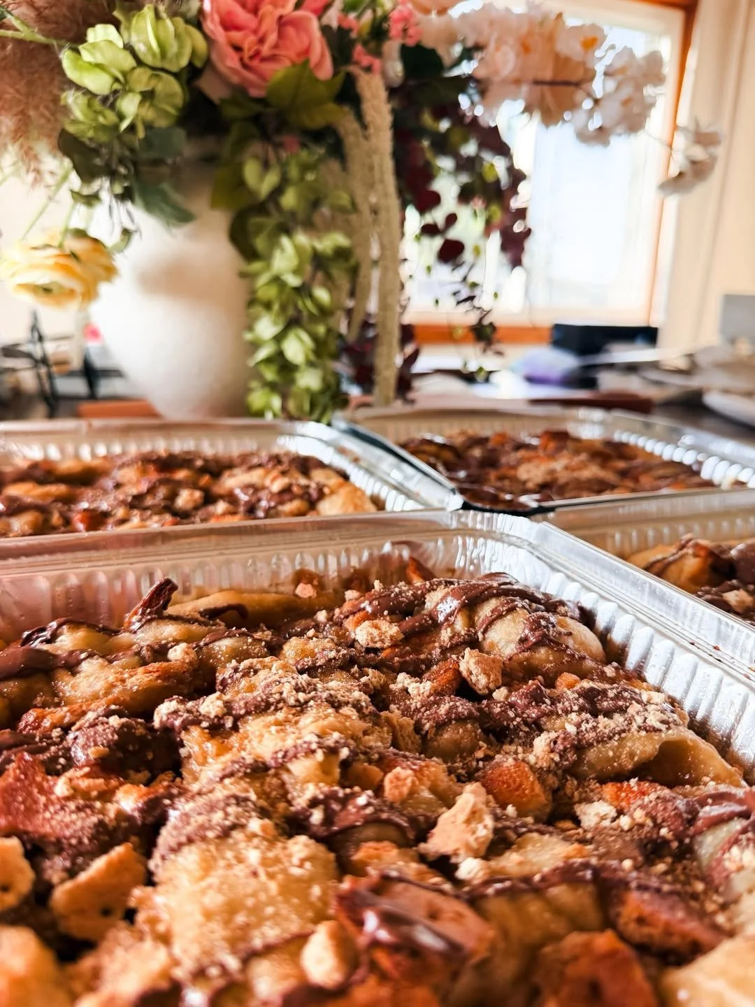 Several trays of baked fruit crisps or cobblers on a table, with a floral arrangement in the background near a window.