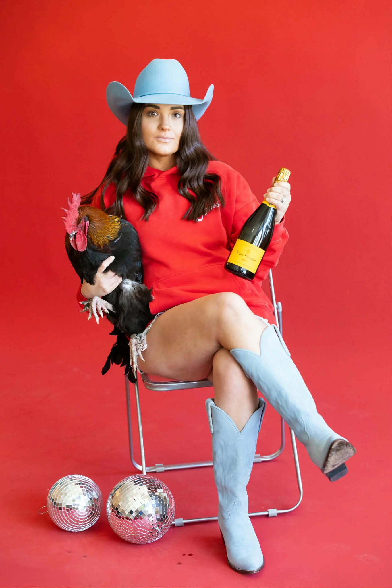 A woman sitting on a chair against a red background, wearing a light blue cowboy hat, a red hoodie, and light blue cowboy boots, holding a small black chicken in her arm and a bottle of champagne in her hand. There are three disco balls on the floor in front of her.