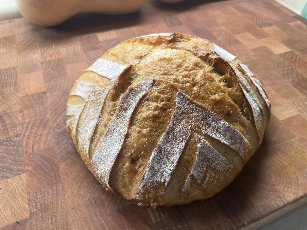 Round loaf of bread with scored crust and flour-dusted surface, resting on a wooden cutting board.
