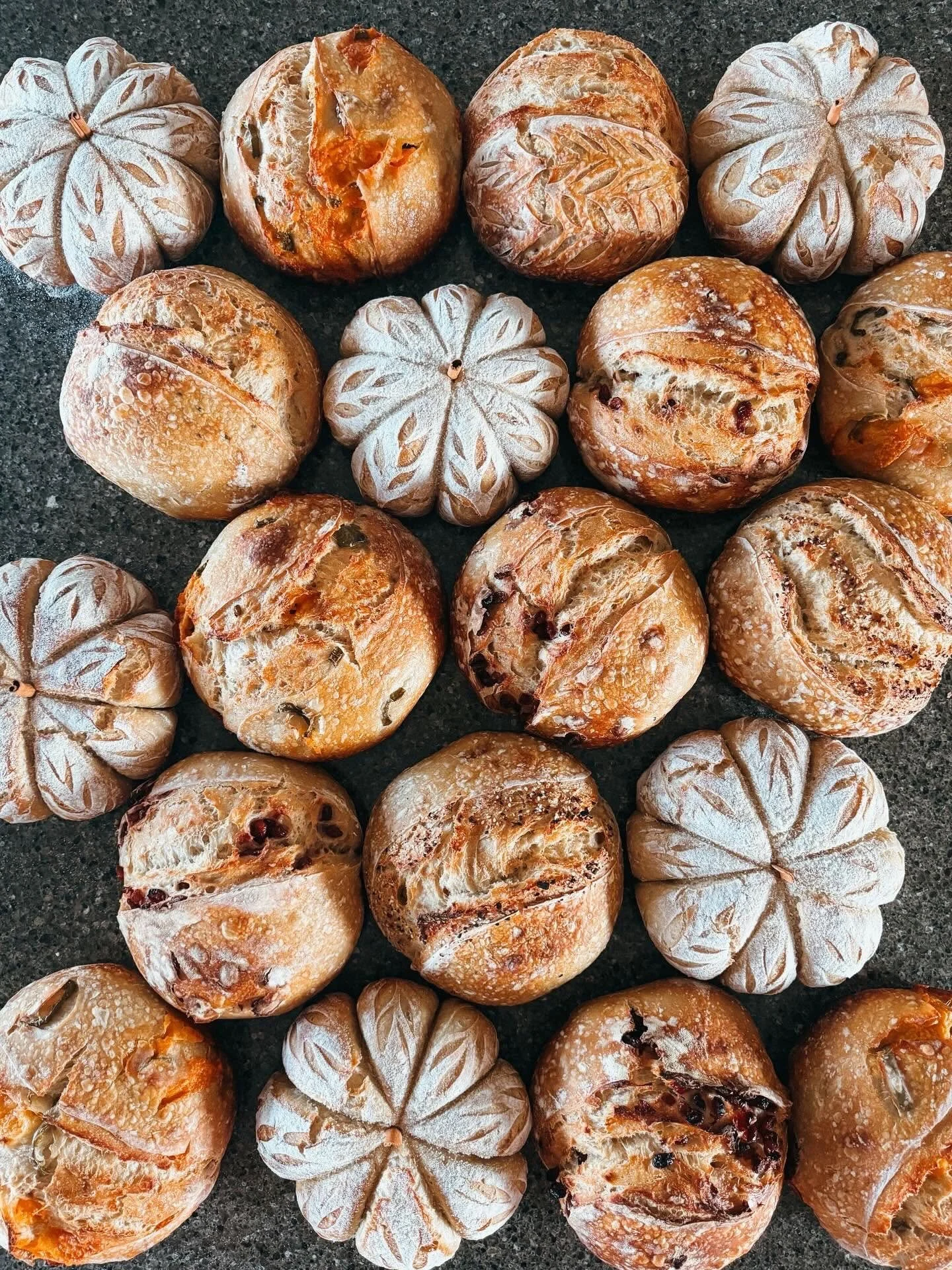 Assorted freshly baked round bread rolls with different textures and toppings, some dusted with flour, arranged on a dark countertop.