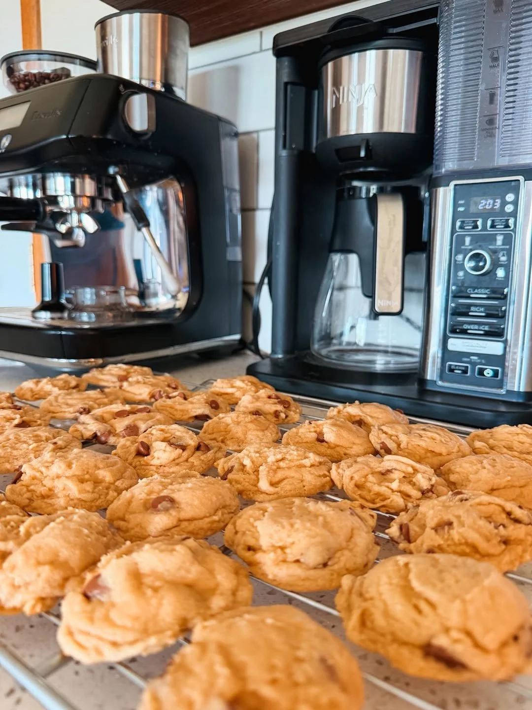 A tray of freshly baked chocolate chip cookies on a cooling rack in a kitchen, with a coffee maker, espresso machine, and a drip coffee maker in the background.