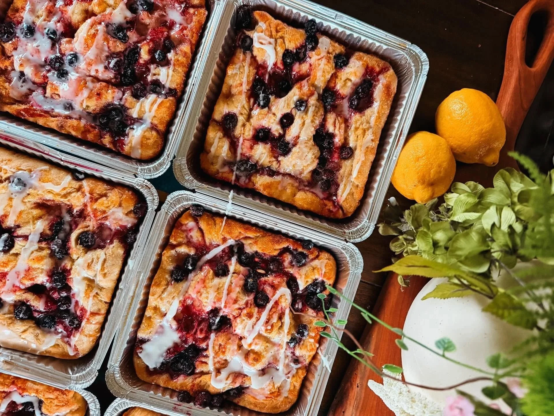 Baked blueberry cheesecake squares in aluminum pans on a dark surface, with lemons, a wooden tray, and green plants nearby.