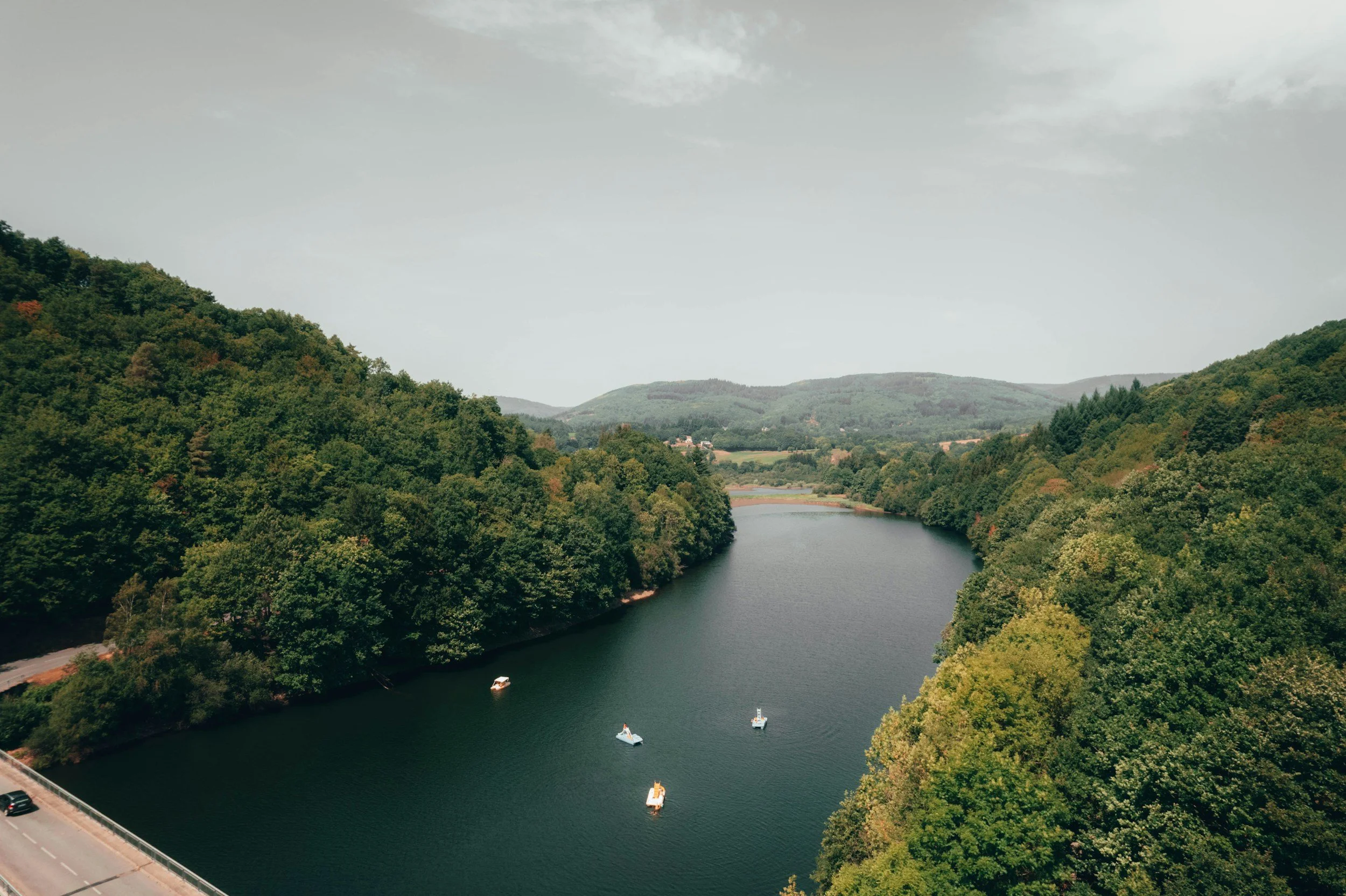 A river winding through a lush green valley with trees on both sides, several boats floating on the water, and hills in the background under a cloudy sky.