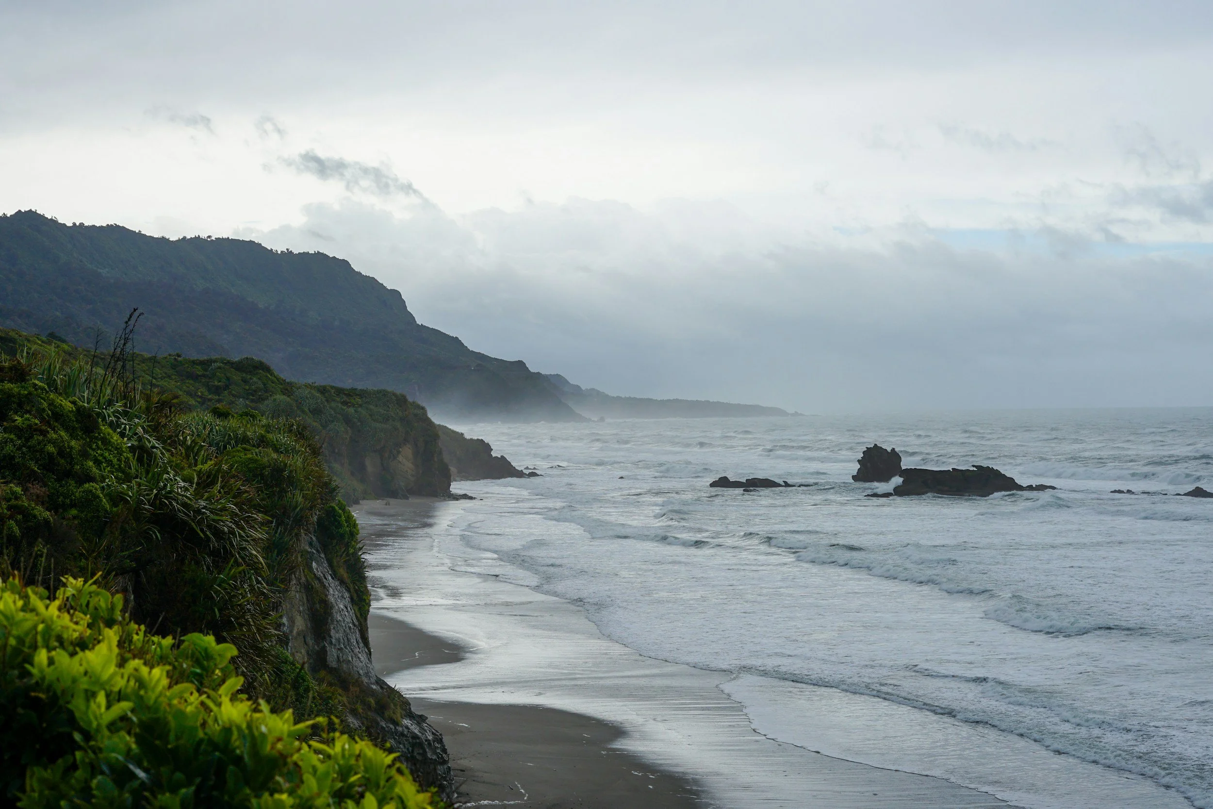 Overcast day on a rugged coastal landscape with green hills, rocky cliffs, and waves crashing onto the sandy beach.