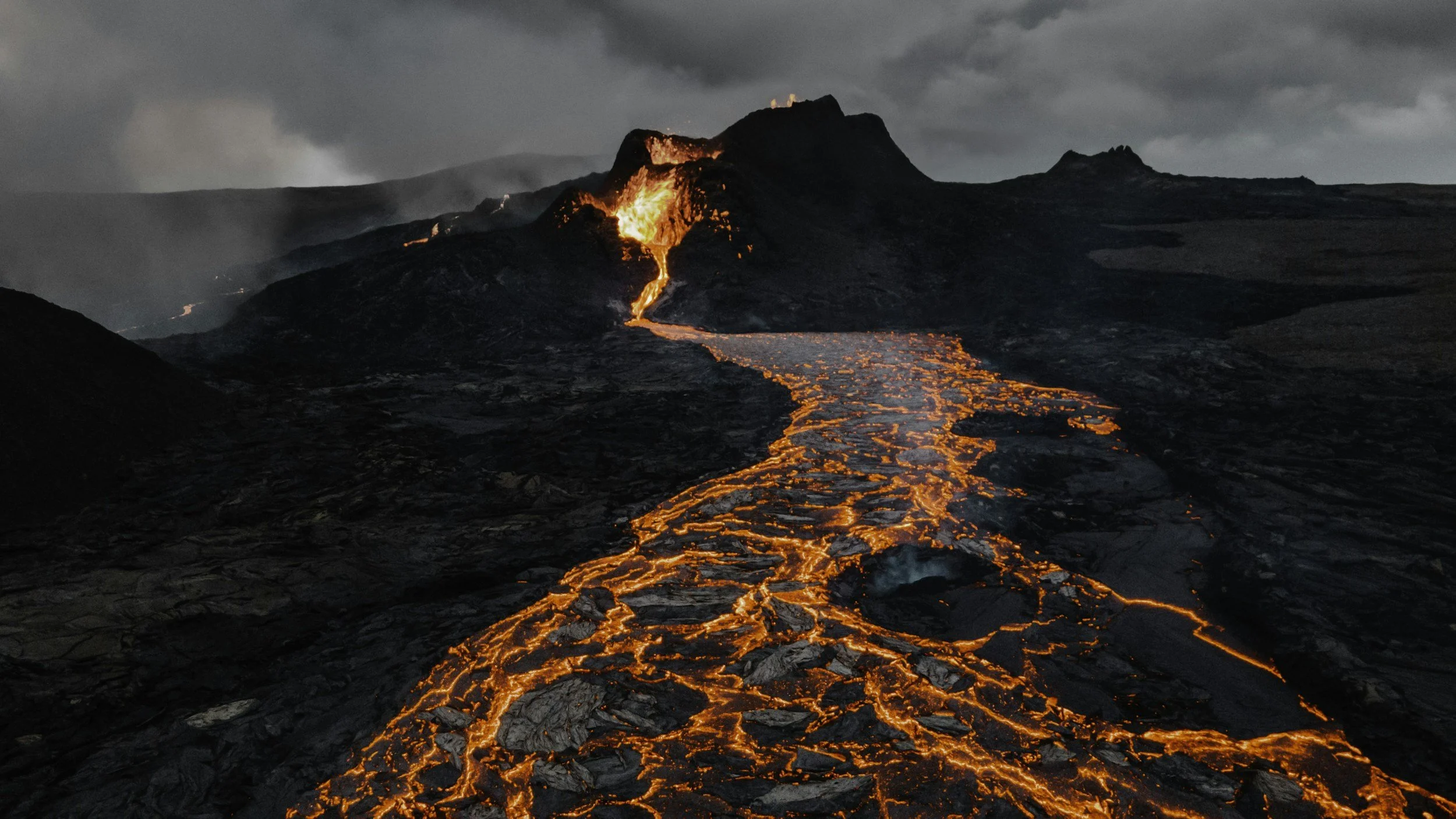 Erupción volcánica activa con lava que fluye y roca volcánica oscura, con cenizas y humo en el cielo.