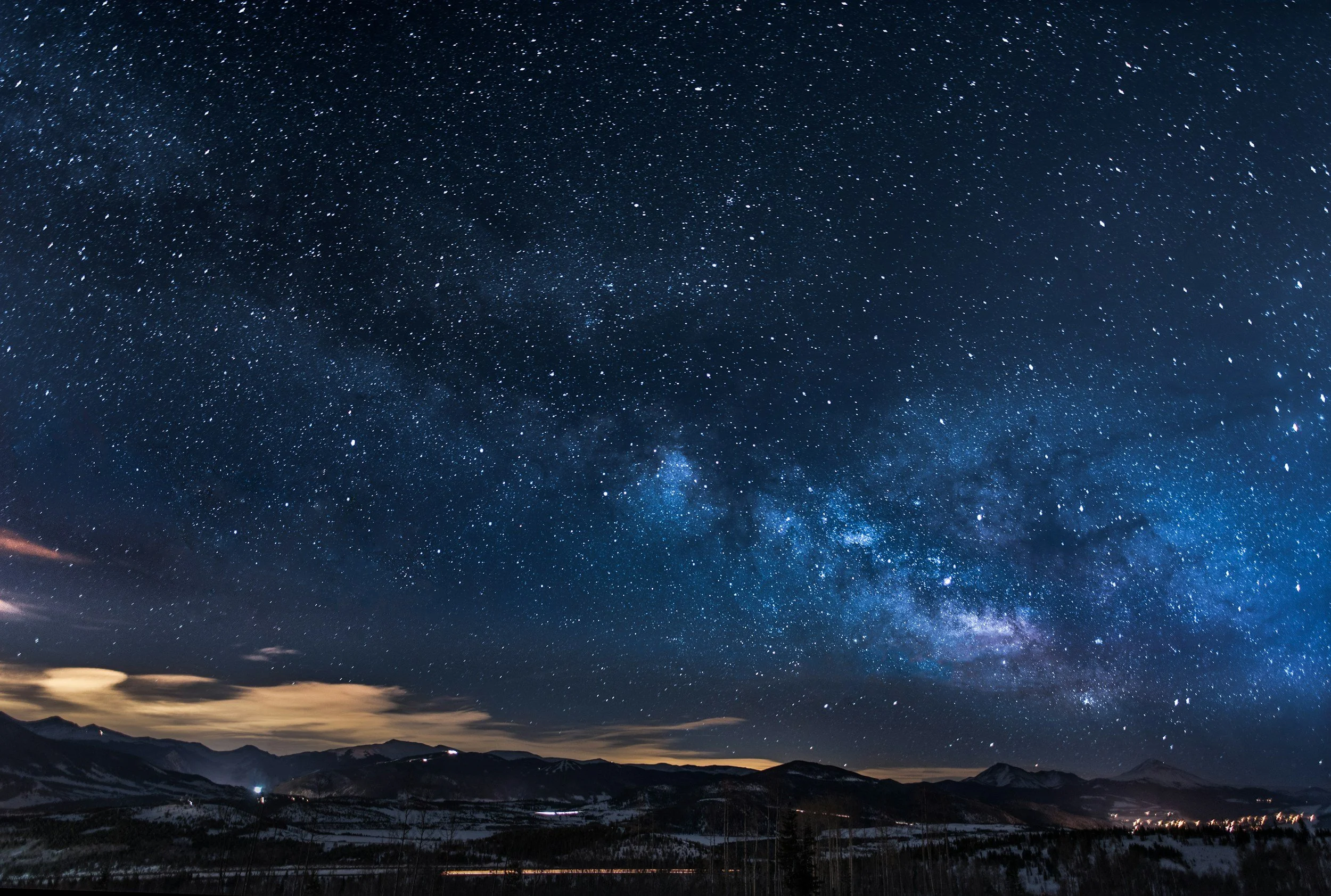 Un cielo nocturno estrellado sobre un paisaje montañoso con algunas nubes cerca del horizonte.