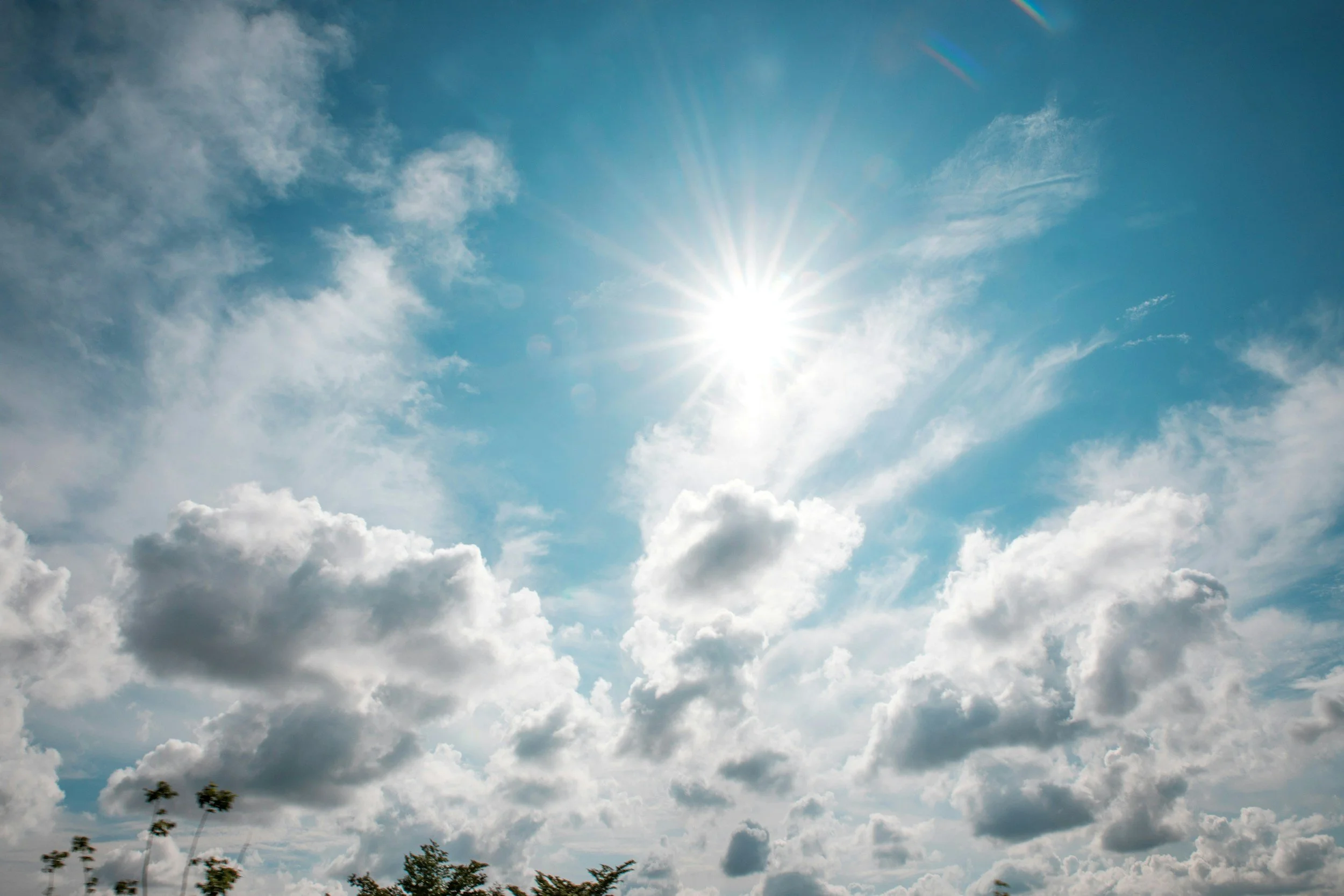 Un sol radiante brilla en un cielo con nubes dispersas, y en la parte inferior de la imagen se divisan algunas copas de árboles.